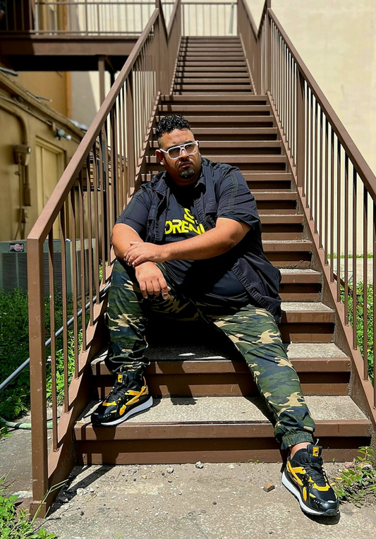 DJ Adrennaa in camo pants and a black shirt sitting on a set of stairs